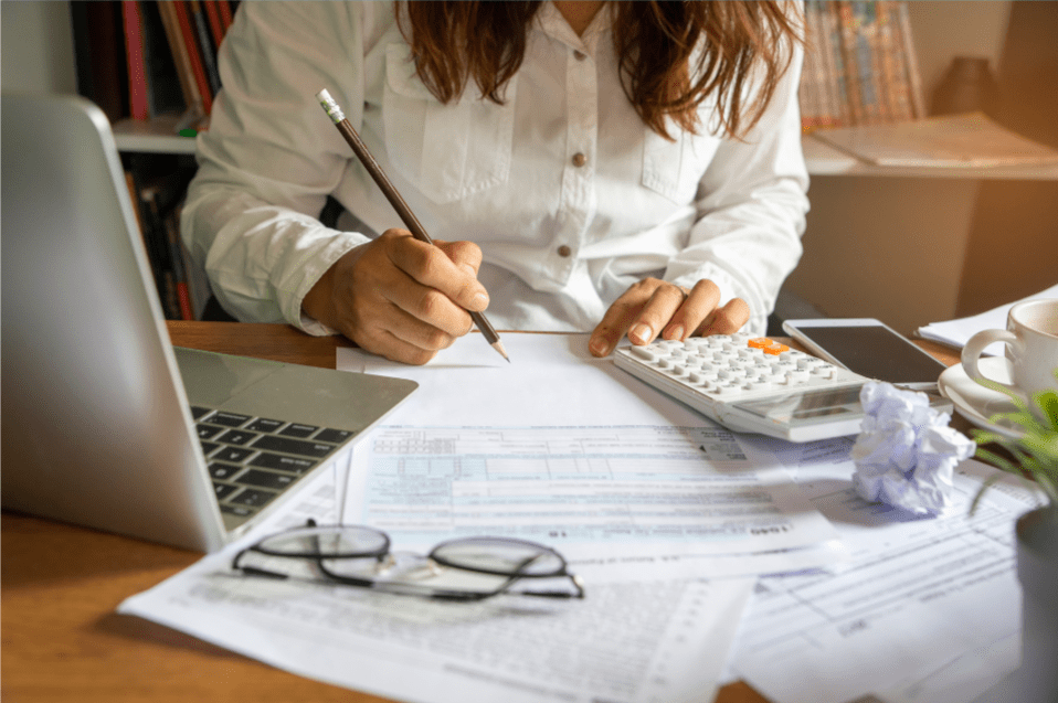 Woman sat at desk with pencil in hand using calculator with paperwork laptop and glasses nearbyk